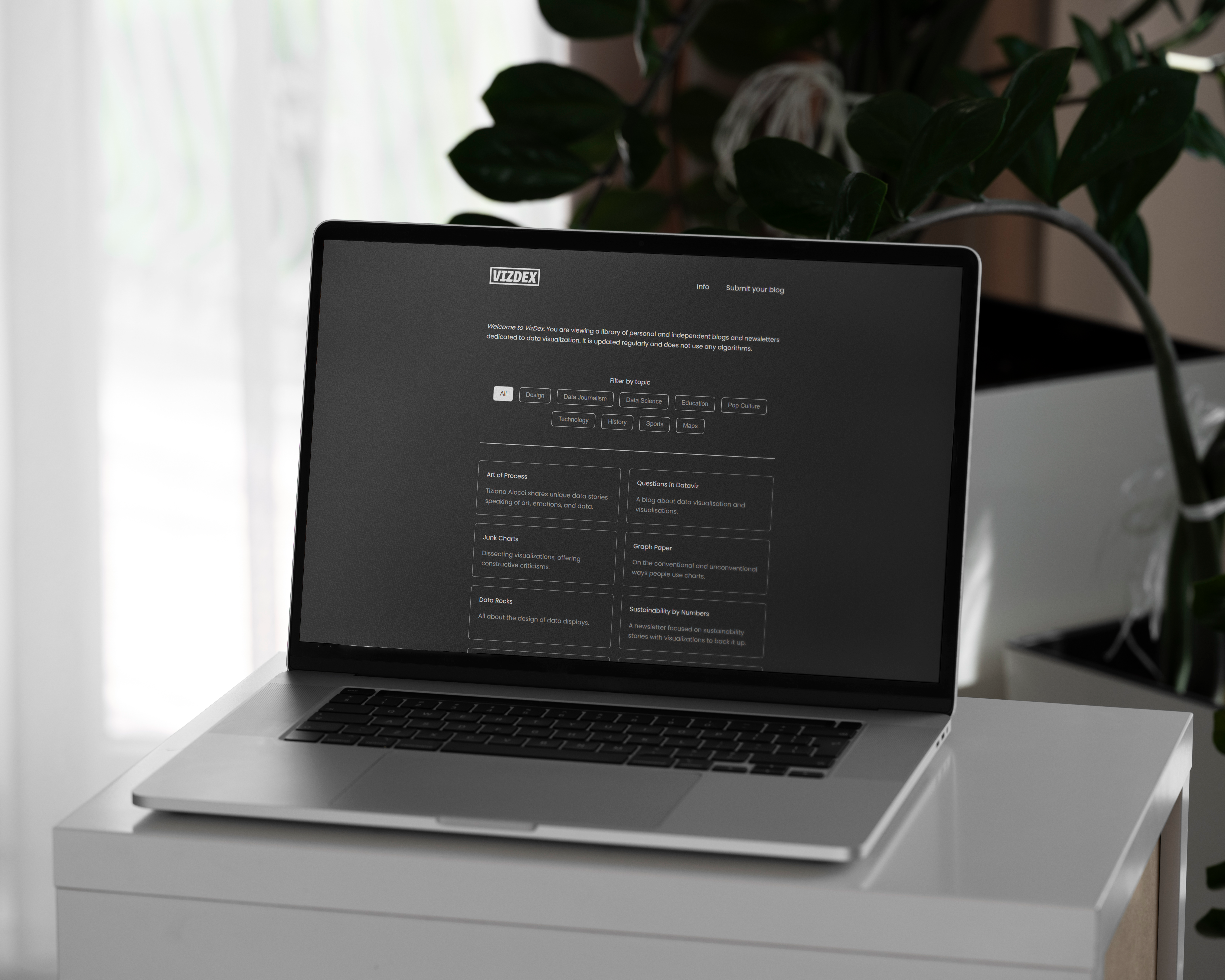 A laptop sitting on a small platform, displaying the homepage of VizDex dot com. The website is dark grey with minimal type.